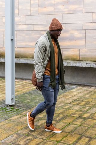 African american man walking plaza wearing beanie, bomber jacket, scarf carrying bag