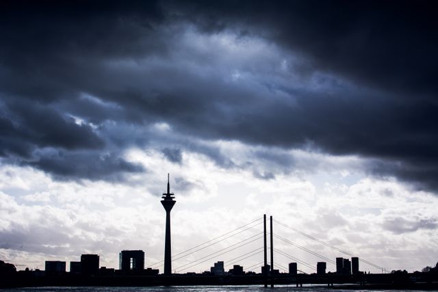 Storm clouds are gathering over a city skyline with a prominent bridge and telecommunication tower visible. The scene features a dramatic sky and an urban landscape, perfect for themes of weather, city life, architecture, or urban planning. This image can be used in articles or advertisements related to travel, real estate, infrastructure, or climate.