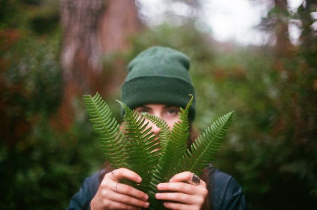 Person wearing green beanie holding fern leaves in the forest, with face partially hidden by leaves. Ideal for use in nature-related content, eco-friendly blogs, outdoor adventure articles, and advertisements promoting environmental conservation.