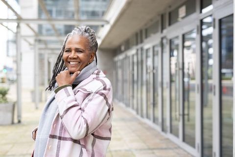 Smiling mature woman standing on urban walkway wearing pink plaid coat and gray scarf