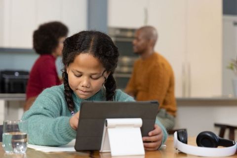 Young girl focusing on tablet for remote learning at kitchen table with parents nearby