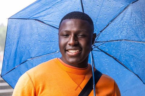 African american man holding blue umbrella smiling in rain wearing orange sweatshirt