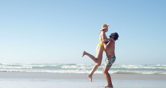 A young Caucasian couple enjoys a playful moment on a sunny beach, with the man carrying the woman in his arms, both smiling and wearing swimwear, with copy space. Their carefree joy and the clear blue sky create a sense of summer bliss and romantic getaway.