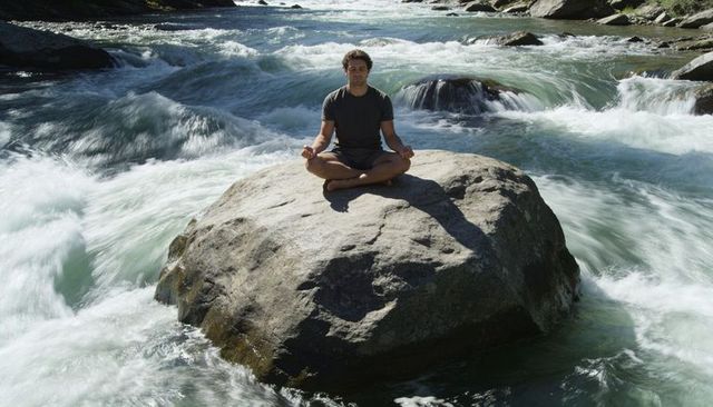 Meditating man sitting cross-legged on boulder amid fast mountain river rapids sunlit