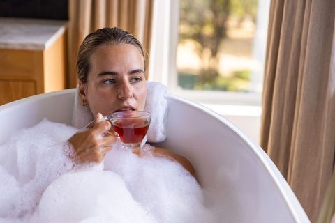 Young woman relaxing in freestanding bubble bath holding glass teacup in sunlit bathroom