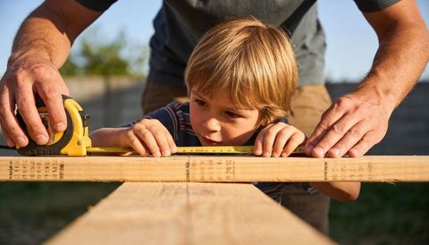 Father guiding son measuring wood with tape measure on backyard diy carpentry project