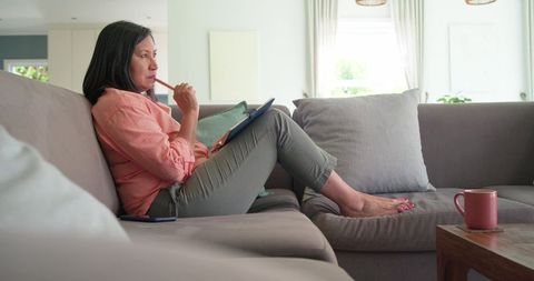 Asian woman jotting notes on sofa with notebook and mug in bright cozy minimalist living room