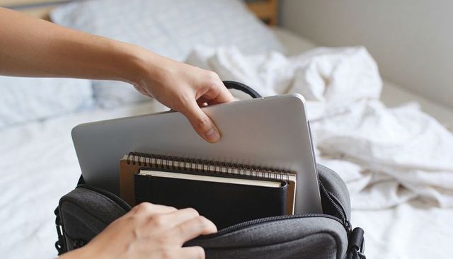 Placing silver laptop and notebooks into gray backpack on bed for minimalist travel