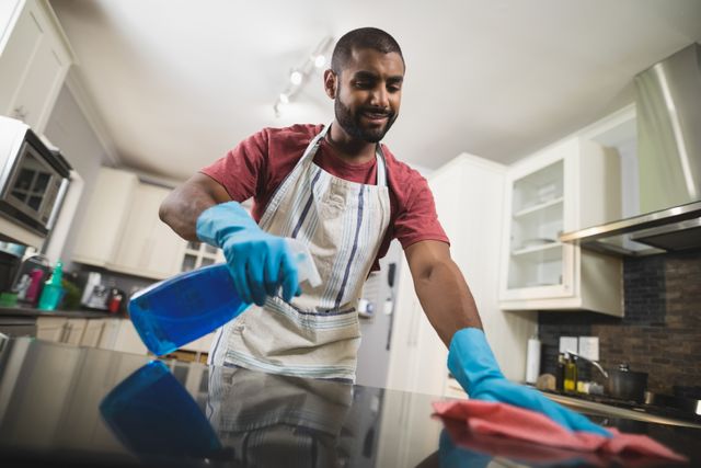 Young man wearing gloves and apron cleaning marble kitchen counter with spray bottle and cloth. Ideal for content related to home cleaning, domestic chores, housekeeping tips, hygiene practices, and modern kitchen maintenance.