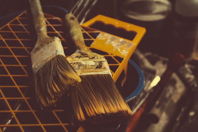 This image captures a close-up view of two well-used paintbrushes resting on a grated surface, surrounded by paint supplies and tools in a workshop. The vintage lighting adds a warm, nostalgic feel to the scene. Ideal for use in articles and advertisements related to painting, home improvement, renovation projects, art and craft supplies, DIY projects, and craftsmanship.