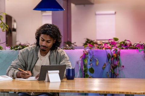 Indian man writing notes while using tablet in modern coworking lounge with plants