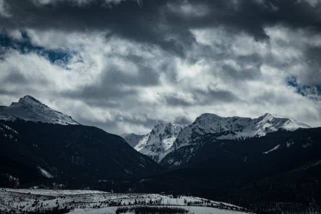 Dramatic image featuring dark clouds hovering over snow-covered mountain peaks, creating a moody and intense atmosphere. Ideal for use in outdoor adventure promotions, travel and tourism campaigns, winter sports advertisements, or illustrations of natural beauty and wilderness exploring.