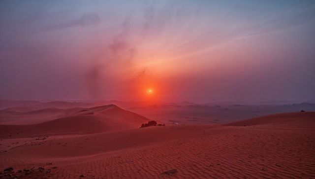 Sunset glowing over red desert dunes with rippled sand patterns and distant rock outcrops
