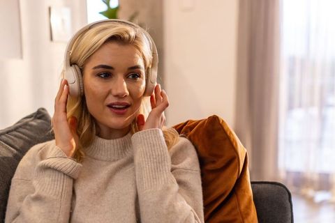 Young woman adjusting wireless headphones while listening on cozy living room sofa
