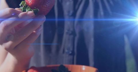 Child holding ripe strawberry over orange bowl with dramatic blue lens flare hand closeup