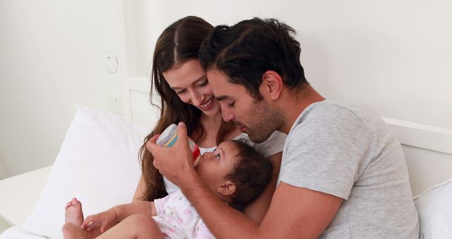 Parents feeding their baby while sitting on a bed in a cozy home. This image can be used for parenting blogs, family care websites, brochures on childcare practices, or advertisements for baby products.