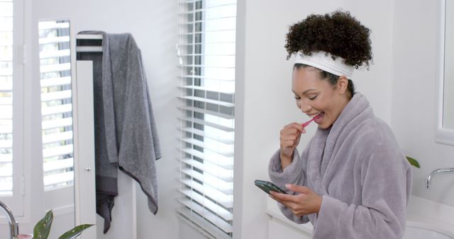 A young woman is multitasking, brushing her teeth while checking her smartphone in a modern, well-lit bathroom. The image highlights a typical busy morning routine combining self-care and technology use. This versatile content can be used for themes related to personal hygiene, modern lifestyle, morning routines, or technology in daily life.