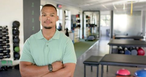 Confident fitness trainer crossing arms in modern gym with smartwatch and free weights
