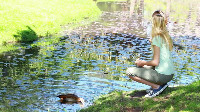 Blonde woman feeding a duck by a tranquil pond with lily pads. Perfect for themes of nature, leisure, outdoor activities, and wildlife. Suitable for blogs, articles, or marketing promoting outdoor experiences, environmental awareness, or calming nature scenes.