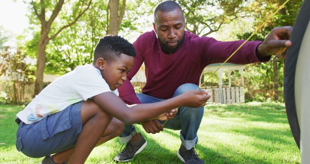 An African American father is showing his young son how to fix a tire in their garden on a bright sunny day. They are both focused on the task. This visual can be used for educational content, family bonding themes, parenting blogs, or highlighting the importance of practical skills and outdoor activities with children.