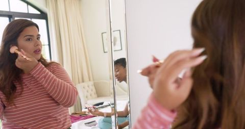 African american friends applying makeup at mirror in bright bedroom while using phone