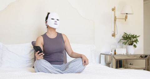 Woman using led facial mask practicing at-home self care routine on bed with smartphone