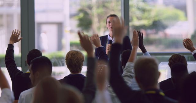 A diverse group of individuals are raising their hands during a business seminar, indicating active participation and engagement in the session. Ideal for illustrating concepts of education, business training, public speaking, interactive learning, and professional development in corporate settings.