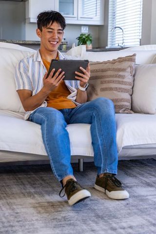 Young asian man relaxing on sofa using tablet and smiling in modern open-plan living room