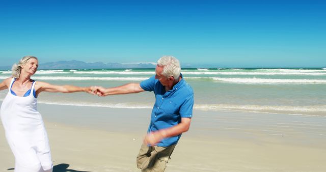 A senior Caucasian couple enjoys a playful moment while holding hands on a sunny beach, with copy space. Their joyful dance against the backdrop of the ocean exemplifies active and happy retirement years.