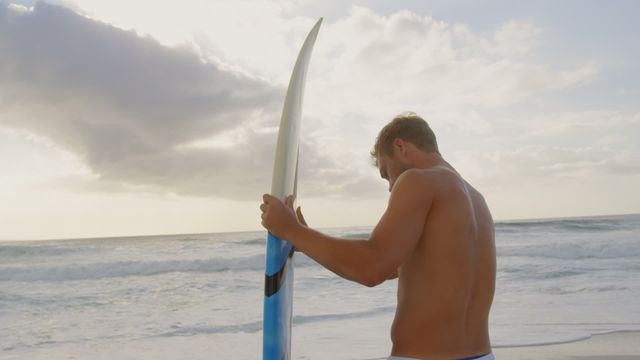 Shirtless young man holding surfboard next to waves at sunset. Ideal for topics on beach activities, summer adventures, coastal living, and water sports. Perfect for lifestyle blogs, travel promotions, and sports advertisement.