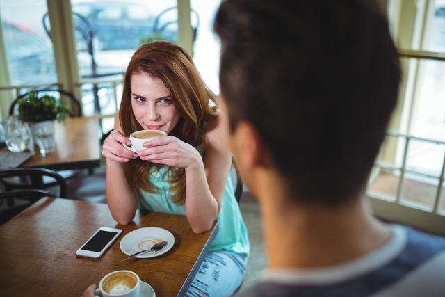Woman enjoying coffee with a friend in a cozy cafe. She is smiling and holding a cup of coffee, while her friend is sitting across the table. Ideal for use in lifestyle blogs, social media posts, advertisements for cafes or coffee brands, and articles about socializing or relaxation.