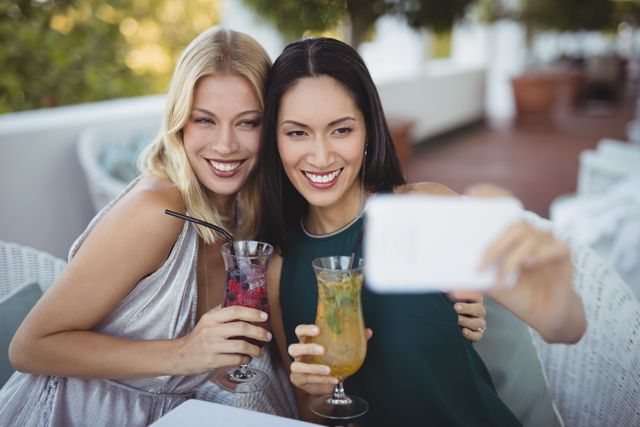 Two women are taking a selfie with a mobile phone at an outdoor restaurant. They are smiling and holding colorful drinks, enjoying a moment of leisure and celebration. This image can be used for themes related to friendship, social media, lifestyle, summer activities, and relaxation.