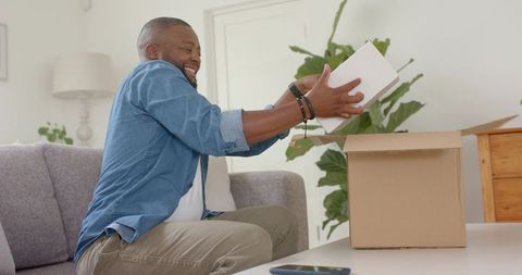 African man happily unboxing delivery in cozy living room with smartphone and houseplant
