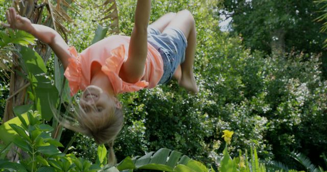 A young Caucasian girl enjoys playtime outdoors, hanging upside down from a tree, surrounded by lush greenery, with copy space. Her joyful expression and the sunny environment convey a sense of carefree childhood fun.
