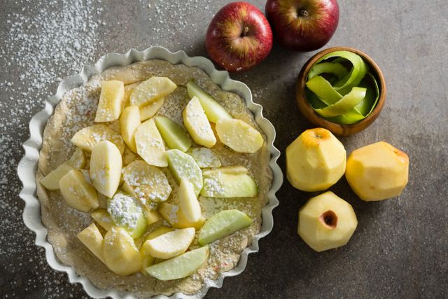 Overhead view of an apple tart in preparation with fresh apple slices and icing sugar. Ideal for use in food blogs, recipe websites, cooking magazines, and culinary tutorials.
