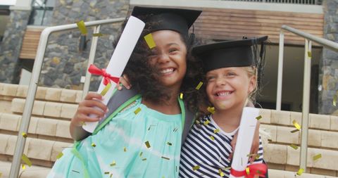 Two young girls standing on stone steps wear graduation caps and hold diplomas, surrounded by gold confetti. Suitable for depicting childhood milestones, academic success, diversity in education, and meaningful friendships formed during school years. Ideal for educational programs, school promotions, and celebratory events.