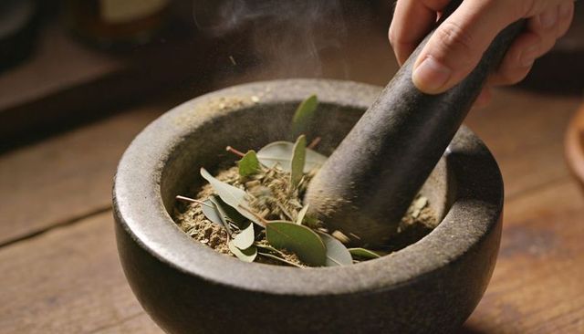 Grinding eucalyptus leaves in stone mortar and pestle releasing aromatic herb dust on wood