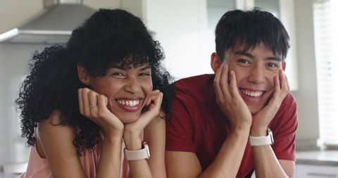 Diverse couple smiling and showing smartwatches while leaning on kitchen counter