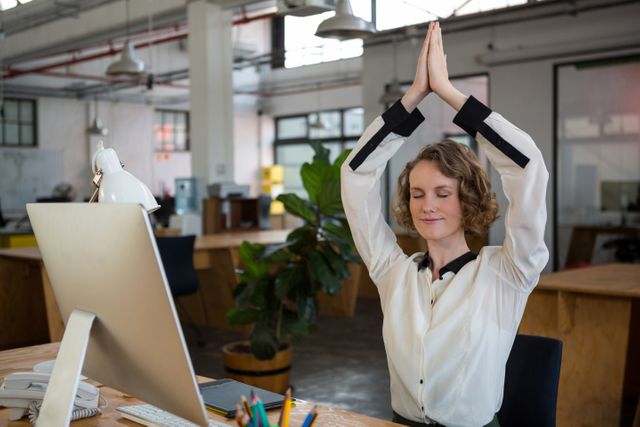 Female graphic designer practicing yoga at her desk in a modern office. Ideal for use in articles or advertisements about workplace wellness, stress relief techniques, mindfulness in the workplace, or promoting healthy lifestyles among professionals.