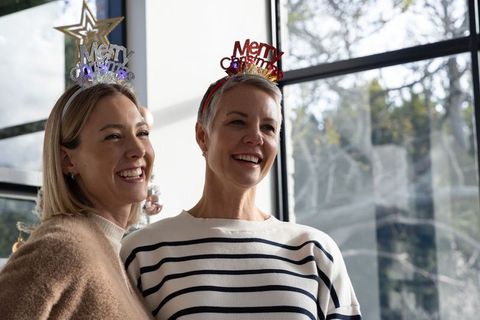 Two women celebrating christmas with festive headbands and cozy sweaters by sunlit window