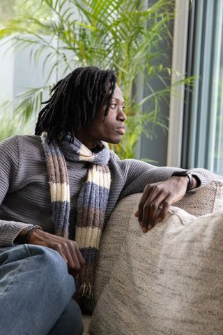 African american man sitting on sofa gazing out window in knit scarf, serene portrait