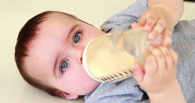 Infant drinking milk from a bottle while lying on a flat surface. Baby is wearing a grey onesie and has striking blue eyes. Great for parenting blogs, baby product advertisements, and nutrition articles.