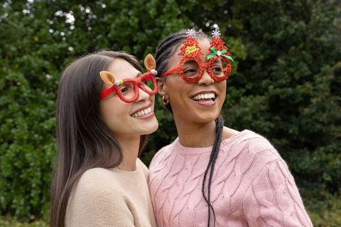 Diverse women wearing festive novelty glasses and knit sweaters smiling outdoors