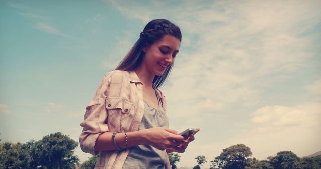 A young Caucasian woman smiles as she looks at her phone, with copy space. Her casual attire and the outdoor setting suggest a relaxed, leisurely day.