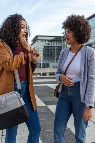 Two women chatting and laughing on urban terrace carrying shoulder bags and wearing blazers