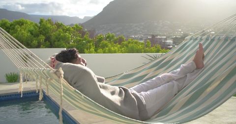Indian man relaxing on striped hammock by pool on sunlit rooftop terrace with mountain city view