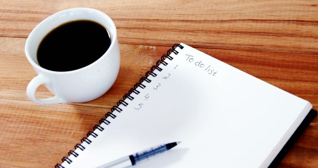 A cup of coffee sits next to a notebook with a to-do list on a wooden table, with copy space. It suggests a moment of planning or organization, at the start of a busy day.