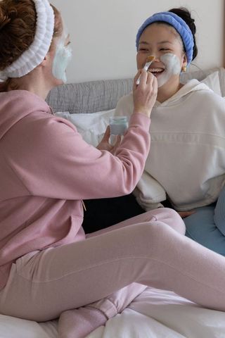 Friends applying blue facial mask on bed during cozy at-home spa ritual