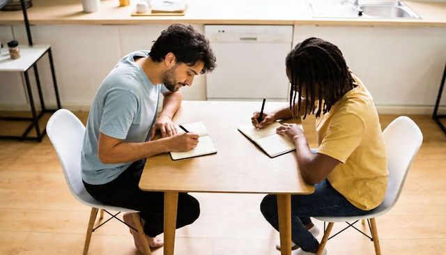 Two men writing at kitchen table collaborating on notebooks in sunlit home workspace