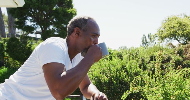 Mature man enjoying coffee outside in garden filled with greenery. Ideal for illustrating concepts of relaxation, healthy morning routines, or enjoying nature at home.